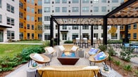 Located at 3025 W Salt Creek Ln in Arlington Heights, Illinois, this thoughtfully designed outdoor courtyard serves as a vibrant communal gathering space nestled within a modern multi-story building complex. The courtyard is anchored by a striking central fire pit lounge area, where a geometric corten steel fire feature with blue glass accents is surrounded by wicker chairs and stylishly patterned side tables, creating an inviting setting for relaxed socializing. A bold black pergola with a warm wood-toned lattice roof frames the lounge area, adding architectural definition and partial shade overhead. Adjacent to the lounge, an outdoor kitchen and bar area features multiple grills and high-top seating, ideal for entertaining. Further into the courtyard, rows of cushioned teak lounge chairs with patio umbrellas offer a serene spot to unwind, overlooking a well-maintained lawn bordered by layered perennial plantings, mature trees, and ornamental shrubs. The paver-lined walkways connect the various zones seamlessly, while the surrounding building facades—finished in a contemporary palette of white and warm amber—enclose the space to create a private, resort-like atmosphere. The overall ambiance is modern, comfortable, and inviting.