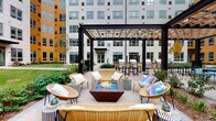 Located at 3025 W Salt Creek Ln in Arlington Heights, Illinois, this thoughtfully designed outdoor courtyard serves as a vibrant communal gathering space nestled within a modern multi-story building complex. The courtyard is anchored by a striking central fire pit lounge area, where a geometric corten steel fire feature with blue glass accents is surrounded by wicker chairs and stylishly patterned side tables, creating an inviting setting for relaxed socializing. A bold black pergola with a warm wood-toned lattice roof frames the lounge area, adding architectural definition and partial shade overhead. Adjacent to the lounge, an outdoor kitchen and bar area features multiple grills and high-top seating, ideal for entertaining. Further into the courtyard, rows of cushioned teak lounge chairs with patio umbrellas offer a serene spot to unwind, overlooking a well-maintained lawn bordered by layered perennial plantings, mature trees, and ornamental shrubs. The paver-lined walkways connect the various zones seamlessly, while the surrounding building facades—finished in a contemporary palette of white and warm amber—enclose the space to create a private, resort-like atmosphere. The overall ambiance is modern, comfortable, and inviting.