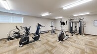 Located at 637 Pine St in Meadville, Pennsylvania, this fitness center offers a well-appointed, single-floor exercise facility designed to support a range of workout routines. The space is finished with neutral-toned carpet tile flooring in a patterned layout and bright fluorescent lighting mounted to the ceiling, creating a clean and energizing environment throughout.

The main exercise area is generously proportioned and houses a variety of cardio equipment, including a treadmill, stationary bikes, and an elliptical machine. A multi-station cable and strength machine by Spirit anchors one corner of the room, complemented by a dedicated free weight rack stocked with a full set of dumbbells. Workout reference charts are posted on the walls to guide users through exercises.

A seating area with two chairs is positioned near a wall-mounted flat-screen television, providing a convenient space to rest or follow along with on-screen content. A wall-mounted split HVAC unit ensures comfortable climate control throughout the space. An adjacent area serves as an entry and transition zone, providing access to an elevator and additional rooms beyond. Natural light filters in through a small window, adding to the overall brightness of the facility. The space is thoughtfully organized, accessible, and equipped to serve a variety of fitness needs.