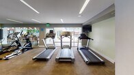 Located at 1718 Ximeno Ave in Long Beach, California, this fitness center offers a well-appointed, single-floor facility designed to support a comprehensive workout experience. The space is finished with a neutral color palette — soft gray and sage green walls paired with durable carpeted flooring — creating a clean, functional environment. Recessed linear LED lighting runs throughout the ceiling, ensuring bright, even illumination across the entire floor.

The cardio zone features multiple treadmills and an elliptical trainer, positioned along a glass wall that opens visually to an outdoor courtyard area, providing natural light and an open, airy feel during workouts. Mounted flat-screen televisions are available for entertainment throughout the space.

The strength training area is generously equipped with a multi-station cable machine, a dedicated weight bench, a leg extension/curl machine, and additional resistance equipment. A full dumbbell rack with a graduated set of free weights rounds out the strength offerings, accommodating a wide range of training needs. Additional cardio options include a stationary cycle and rowing-style equipment.

The layout is thoughtfully organized to allow ease of movement between stations. Windows along multiple walls bring in natural light and views of the surrounding area. Overall, this fitness center provides a modern, well-equipped environment suited for both cardio and strength training routines.