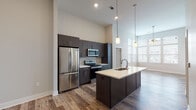 This contemporary single-floor residence showcases a thoughtfully designed open-concept layout that seamlessly connects the kitchen and living areas. The kitchen is a standout feature, equipped with stainless steel appliances including a French door refrigerator, electric range, and built-in microwave. Dark espresso cabinetry extending to the ceiling provides ample storage, while a large center island with a light quartz countertop and undermount sink serves as the focal point of the space. Stylish pendant lighting illuminates the island, complementing the soaring ceilings that lend a sense of grandeur throughout. Warm hardwood-style flooring flows through the main living areas and entryway, transitioning to plush carpet in the bedrooms for added comfort. The living areas are bathed in natural light through expansive windows dressed with horizontal blinds, offering views of the surrounding neighborhood. Ceiling fans are present in the living and bedroom spaces for added climate control. Bedrooms feature neutral-toned walls and are generously proportioned. The bathroom includes wood-look tile flooring and clean, modern fixtures. A dedicated entryway corridor provides a welcoming transition into the home.