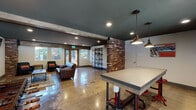 Located at 845 Hancock Street in Hayward, CA, this single-floor community space combines industrial and modern design elements to create a functional and visually striking environment. The interior is defined by polished concrete floors that reflect natural and recessed lighting throughout, contributing to the open, loft-like atmosphere. A deep teal ceiling pairs with light gray walls to establish a bold yet cohesive color palette.

Exposed brick columns serve as prominent architectural focal points, adding warmth and texture against the otherwise modern finishes. Structural beams with a reclaimed wood aesthetic reinforce the industrial character of the space. The lounge area features comfortable seating, a wall-mounted flat-screen television, and direct access to the outdoors through wood-framed glass doors that invite natural light and garden views.

A dedicated recreation zone includes a foosball table positioned near an exterior door, while an open-concept kitchen offers white cabinetry, dark countertops, stainless steel appliances, and a window overlooking the exterior. Industrial-style pendant lights and open metal shelving units complete the aesthetic. A wall-mounted mini-split unit provides climate control, and multiple exterior access points connect the interior to surrounding outdoor areas.