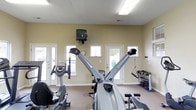 This single-floor fitness room offers a functional and well-equipped workout space designed to support a variety of exercise routines. The room features light yellow walls, a neutral carpeted floor, and a textured ceiling with overhead fluorescent lighting, creating a clean and practical environment. Natural light filters in through multiple white-framed windows positioned along the walls, complementing the bright, open feel of the space. Two exterior doors provide convenient access points, further enhancing ventilation and ease of entry.

The fitness area is outfitted with a selection of cardio machines, including a recumbent stationary bike and what appears to be a rowing or dual-action exercise machine positioned prominently in the center of the room. A wall-mounted television is installed above the windows, allowing users to watch content while exercising. Along one side of the room, a dumbbell rack holds a range of free weights, accompanied by additional strength training accessories, offering options for resistance-based workouts. The overall layout is straightforward and accessible, making efficient use of the available floor space while keeping equipment organized and easy to navigate.