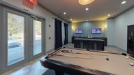 Located at 6310 Ansley Falls Dr in Charlotte, NC, this single-floor community amenity space is designed for social gathering and recreation. The room centers around a full-size billiards table positioned on a patterned area rug over polished tile flooring, providing ample space for play and movement. Recessed lighting and a pendant drum fixture illuminate the room evenly, contributing to a relaxed yet lively atmosphere.

Along one wall, a built-in bar counter with dark granite surfaces is flanked by bar stools and backed by a mosaic tile accent wall. Two wall-mounted flat-screen televisions are positioned above the bar, making this area ideal for watching sports or entertainment while socializing. A glass-front dark wood bookcase offers a small library of reading materials for guests.

The space is finished with soft gray walls, crown molding, and navy blue grommet-panel curtains that frame the glass panel doors leading to an outdoor area. A cozy seating nook with a upholstered settee and accent pillows is anchored beneath a large abstract canvas painting, adding an artistic focal point. The combination of entertainment amenities, comfortable seating, and outdoor connectivity makes this a well-rounded communal gathering space.