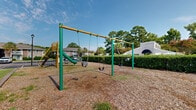 Located at 200 Branchwood Dr in Goose Creek, South Carolina, this outdoor community space serves as a dedicated recreational area for residents. The playground is the central feature, equipped with a multi-seat swing set framed in green and yellow metal and a standalone slide structure with an attached climbing ladder. The play surface is covered with wood chip mulch, providing a safety-conscious ground covering throughout the area. The playground is neatly bordered by well-maintained hedgerows that define the space and offer a sense of enclosure and privacy. Mature trees, including tall pines and palms, surround the broader community, providing natural shade and a lush backdrop under the open South Carolina sky. Adjacent to the playground, the community includes ample paved parking, open grassy areas, and additional outdoor gathering spaces. A community building with a distinctive peaked roofline and arched accent is visible nearby, suggesting shared amenity facilities on the grounds. The overall setting reflects a well-established residential community with thoughtfully maintained outdoor spaces designed to support an active, community-oriented lifestyle.