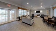 Located at 168 River Rd in Andover, Massachusetts, this single-floor community amenity space is thoughtfully designed to provide residents with a comfortable and functional gathering environment. The main lounge area is anchored by a classic white-mantel fireplace with a wall-mounted television, creating a warm focal point for relaxation and socializing. Plush sectional seating and accent chairs are arranged to encourage conversation, while recessed lighting and elegant crown molding lend the space a refined, polished character. Large windows dressed with neutral blinds and gold valances allow natural light to fill the room, and a French door provides direct access to the outdoors. The carpeted lounge flows naturally into a dining area furnished with a full-sized table and chairs, making the space well-suited for community gatherings or events. Adjacent to the living and dining areas, a well-appointed kitchen features white raised-panel cabinetry, granite countertops, tile flooring, and stainless steel appliances including a refrigerator, wall oven, and microwave. The overall aesthetic is cohesive and inviting, blending classic architectural details with modern conveniences to create a versatile, community-focused environment.