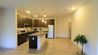 This single-floor residence offers a thoughtfully designed open-concept layout that seamlessly connects the kitchen and living areas, creating a spacious and inviting atmosphere. The kitchen is a standout feature, showcasing rich dark-espresso cabinetry that extends to the upper walls, providing generous storage space. A granite-topped center island serves as both a functional workspace and a casual dining spot, while pendant lighting above adds a stylish focal point. Stainless steel appliances, including a range, microwave, and refrigerator, lend a modern, professional feel to the space. Light-toned, hardwood-style flooring runs throughout the open area, complementing the warm, neutral wall tones and contributing to a bright, cohesive aesthetic. Recessed ceiling lighting ensures the space is well-lit throughout the day, while the open sightlines from the kitchen into the living area make the floor plan feel expansive and connected. The overall ambiance is clean, contemporary, and functional — well-suited for both everyday living and entertaining.