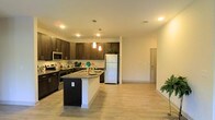 This single-floor residence offers a thoughtfully designed open-concept layout that seamlessly connects the kitchen and living areas, creating a spacious and inviting atmosphere. The kitchen is a standout feature, showcasing rich dark-espresso cabinetry that extends to the upper walls, providing generous storage space. A granite-topped center island serves as both a functional workspace and a casual dining spot, while pendant lighting above adds a stylish focal point. Stainless steel appliances, including a range, microwave, and refrigerator, lend a modern, professional feel to the space. Light-toned, hardwood-style flooring runs throughout the open area, complementing the warm, neutral wall tones and contributing to a bright, cohesive aesthetic. Recessed ceiling lighting ensures the space is well-lit throughout the day, while the open sightlines from the kitchen into the living area make the floor plan feel expansive and connected. The overall ambiance is clean, contemporary, and functional — well-suited for both everyday living and entertaining.