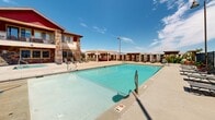 Located in Draper, Utah, this outdoor recreational amenity area is thoughtfully designed to provide a resort-style experience against a backdrop of breathtaking Wasatch Mountain views. The centerpiece is a generously sized swimming pool with crystal-clear turquoise water, offering both shallow and deeper sections to accommodate a range of swimmers. A rinse station is conveniently positioned poolside, and safety equipment is readily accessible throughout the space.

The pool deck is finished with a durable, textured concrete surface and is surrounded by ample open space for sunbathing and relaxation. Lounge chairs and seating are arranged along the perimeter, allowing guests to enjoy the abundant Utah sunshine. A row of additional poolside chairs lines a masonry retaining wall, maximizing the use of every corner of the area.

The complex is enclosed by ornamental iron fencing with a secure, self-closing access gate, complemented by striking natural stone columns that lend an upscale architectural character to the space. A pergola structure adds a shaded gathering area, while decorative pole lighting provides illumination for evening use.

Adjacent to the pool, a children's playground with a colorful multi-level play structure and slide offers family-friendly entertainment. The open-air setting, framed by panoramic mountain scenery and clear blue skies, creates an inviting and vibrant outdoor environment.