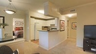 This single-floor residence offers a thoughtfully designed layout that blends functionality with a welcoming ambiance. The kitchen serves as the heart of the space, featuring crisp white cabinetry, a prominent overhead range hood, and a peninsula with a dark countertop that doubles as a casual dining or prep area. A full-size refrigerator and integrated appliances complete the kitchen's practical setup. The open-concept design allows the kitchen to flow seamlessly into the adjacent living area, which is finished with warm, neutral-toned carpeting that adds comfort underfoot. Light-toned walls throughout create a bright, airy feel, complemented by recessed and flush-mount ceiling lighting that ensures the space is well-lit. A hallway extending from the main living area leads to additional rooms, with framed wall art adding tasteful decorative touches along the way. The transition between the kitchen's hard flooring and the living area's carpeting subtly defines each zone while maintaining visual continuity. Overall, the space presents a clean, neutral palette that is both inviting and versatile, offering a solid foundation for personalization.