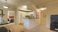 This single-floor residence offers a thoughtfully designed layout that blends functionality with a welcoming ambiance. The kitchen serves as the heart of the space, featuring crisp white cabinetry, a prominent overhead range hood, and a peninsula with a dark countertop that doubles as a casual dining or prep area. A full-size refrigerator and integrated appliances complete the kitchen's practical setup. The open-concept design allows the kitchen to flow seamlessly into the adjacent living area, which is finished with warm, neutral-toned carpeting that adds comfort underfoot. Light-toned walls throughout create a bright, airy feel, complemented by recessed and flush-mount ceiling lighting that ensures the space is well-lit. A hallway extending from the main living area leads to additional rooms, with framed wall art adding tasteful decorative touches along the way. The transition between the kitchen's hard flooring and the living area's carpeting subtly defines each zone while maintaining visual continuity. Overall, the space presents a clean, neutral palette that is both inviting and versatile, offering a solid foundation for personalization.