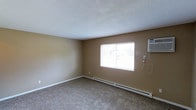 This single-floor unit features two bedrooms and one bathroom within a well-proportioned floor plan. The living area, pictured here, showcases neutral beige walls, wall-to-wall carpet flooring, and a textured ceiling that contributes to the room's understated character. A centrally positioned window allows natural light to filter in, offering a view of the surrounding outdoor greenery. Climate control is handled by a wall-mounted air conditioning unit and a baseboard heating system running along the exterior wall, ensuring year-round comfort. Clean white baseboards provide a crisp contrast to the warm-toned walls throughout. The layout is straightforward and functional, offering residents flexibility in how they arrange and utilize the space. The unit's single-floor design ensures easy accessibility and a cohesive flow between rooms. Overall, the space presents a clean, move-in-ready environment with practical features that cater to comfortable residential living.