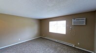 This single-floor unit features two bedrooms and one bathroom within a well-proportioned floor plan. The living area, pictured here, showcases neutral beige walls, wall-to-wall carpet flooring, and a textured ceiling that contributes to the room's understated character. A centrally positioned window allows natural light to filter in, offering a view of the surrounding outdoor greenery. Climate control is handled by a wall-mounted air conditioning unit and a baseboard heating system running along the exterior wall, ensuring year-round comfort. Clean white baseboards provide a crisp contrast to the warm-toned walls throughout. The layout is straightforward and functional, offering residents flexibility in how they arrange and utilize the space. The unit's single-floor design ensures easy accessibility and a cohesive flow between rooms. Overall, the space presents a clean, move-in-ready environment with practical features that cater to comfortable residential living.