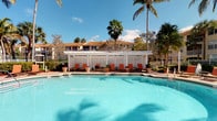 This single-level outdoor amenity space centers around a generously sized swimming pool with crystal-clear turquoise water, offering a true resort-like atmosphere. The pool is complemented by a striking white pergola structure along one side, which provides a shaded retreat from the sun. Beneath the pergola, a row of vibrant orange lounge chairs and side tables are neatly arranged, creating a comfortable and inviting relaxation area. Decorative potted plants are placed throughout, adding color and a touch of natural warmth to the space. The surrounding landscape features mature palm trees and lush tropical greenery that frame the area beautifully against a bright blue sky. In the background, a multi-story residential building with a warm terra cotta roof is visible, reinforcing the cohesive architectural character of the community. A white lattice privacy screen borders the pergola, adding both aesthetic detail and a sense of enclosure. The overall ambiance is bright, airy, and tropical, making this an ideal outdoor gathering and leisure destination designed for comfort and relaxation in a sun-drenched setting.