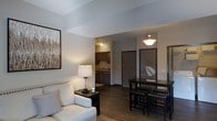This single-floor residence offers a thoughtfully designed open-concept layout that integrates the living, dining, and kitchen areas into one cohesive and functional space. The living area is anchored by a comfortable upholstered sofa and complemented by warm accent lighting, creating an inviting atmosphere for relaxation. A framed piece of wall art adds a decorative touch to the neutral-toned walls. Adjacent to the living area, a dark-finished dining table with bar-height seating provides a stylish space for meals and gatherings. The kitchen, visible through the open layout, features dark cabinetry, a sink with overhead lighting, and practical counter space, offering an efficient cooking environment. Rich, dark hardwood-style flooring flows consistently throughout the space, adding warmth and visual continuity. The entryway, framed by a dark door, is neatly integrated into the overall layout. Angled ceiling details add an architectural dimension to the space, enhancing its modern character. Pendant and overhead lighting fixtures provide layered illumination, creating a well-lit yet comfortable ambiance throughout the unit. Overall, the space balances contemporary design with everyday functionality.