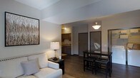 This single-floor residence offers a thoughtfully designed open-concept layout that integrates the living, dining, and kitchen areas into one cohesive and functional space. The living area is anchored by a comfortable upholstered sofa and complemented by warm accent lighting, creating an inviting atmosphere for relaxation. A framed piece of wall art adds a decorative touch to the neutral-toned walls. Adjacent to the living area, a dark-finished dining table with bar-height seating provides a stylish space for meals and gatherings. The kitchen, visible through the open layout, features dark cabinetry, a sink with overhead lighting, and practical counter space, offering an efficient cooking environment. Rich, dark hardwood-style flooring flows consistently throughout the space, adding warmth and visual continuity. The entryway, framed by a dark door, is neatly integrated into the overall layout. Angled ceiling details add an architectural dimension to the space, enhancing its modern character. Pendant and overhead lighting fixtures provide layered illumination, creating a well-lit yet comfortable ambiance throughout the unit. Overall, the space balances contemporary design with everyday functionality.