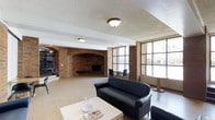 This single-floor lounge space is defined by its prominent architectural character, centered around a large arched brick fireplace that serves as the focal point of the room. The surrounding exposed brick walls extend throughout, lending a warm, rustic aesthetic that contrasts with the textured white ceiling above. Flush-mount ceiling fixtures provide consistent overhead lighting throughout the space.

The layout is open and generously sized, accommodating multiple seating arrangements. Black upholstered sofas and chairs are positioned around a light wood coffee table, creating a comfortable gathering area. A separate seating zone near the large multi-pane windows offers additional space for smaller groups or individuals.

The oversized windows, framed in black metal, line one wall and allow considerable natural light to filter into the interior, enhancing the overall brightness of the space. The flooring transitions between tile and hardwood-style surfaces, reinforcing the functional yet inviting atmosphere.

Overall, the space combines classic brick craftsmanship with practical furnishings, making it well-suited as a shared lounge or common area for residents or visitors.