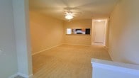 This single-floor residence offers a straightforward and functional layout designed for comfortable everyday living. The main living area is generously proportioned, featuring warm beige carpeting that extends throughout the space and neutral-toned walls that create a bright, welcoming ambiance. A ceiling fan with an integrated light fixture is centrally positioned overhead, providing both air circulation and ambient lighting.

A pass-through counter connects the living area to the kitchen, creating an open, connected feel between the two spaces. The kitchen is equipped with dark appliances, including a range and refrigerator, and features wood-toned cabinetry that adds warmth to the space. A window above the kitchen counter allows natural light to filter in.

Adjacent to the kitchen, a hallway leads to the unit's entry door, which is fitted with a secure white paneled door, adding a clean, classic finish to the entryway. The overall color palette throughout the space is cohesive and understated, with warm neutral tones that lend themselves well to a variety of interior styles. The open flow between the living and kitchen areas makes the space feel practical and easy to navigate, suitable for both everyday use and casual entertaining.