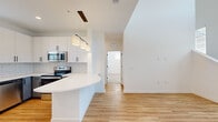 This contemporary two-story home is designed with a clean, modern aesthetic and an open, airy atmosphere. The main living area impresses with dramatically high ceilings and large windows that flood the space with natural light, creating a bright and welcoming environment. Light-toned hardwood-style flooring extends throughout the main level, complementing the crisp white walls and adding warmth to the open layout.

The kitchen is a standout feature, showcasing a thoughtful two-tone cabinet design with white upper cabinets and grey lower cabinets, all accented by sleek matte black hardware. A classic white subway tile backsplash lines the walls, while a curved white quartz peninsula provides additional prep and seating space. Stainless steel appliances, including a range, microwave, refrigerator, and dishwasher, complete the well-appointed kitchen. A trio of pendant lights on a black rail fixture adds a stylish focal point above the peninsula.

Bedrooms are carpeted for comfort and benefit from ample natural light through generously sized windows. One bedroom features direct outdoor access through a glass-panel door. The bathroom is finished with a grey vanity, matte black fixtures, a walk-in glass-enclosed shower with white tile surround, and wood-look tile flooring. A staircase connects the two floors, and built-in closet shelving provides practical storage solutions throughout.