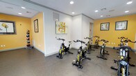 This single-floor spin studio is thoughtfully designed to accommodate focused cycling workouts in a clean, energizing environment. The room features several stationary spin bikes arranged in a structured layout, providing ample space for each user to exercise comfortably without feeling crowded.

The interior design strikes a balance between energizing and functional. Bold yellow accent walls contrast with crisp white trim and neutral upper walls, creating a motivating atmosphere suited for high-intensity workouts. Recessed ceiling lights provide consistent, even illumination throughout the space, ensuring a well-lit environment at all times.

The flooring consists of durable, speckled gray rubberized material, a practical choice that offers traction, noise reduction, and easy maintenance. Decorative framed artwork adorns the walls, adding a touch of visual interest without distracting from the room's primary purpose.

A doorway visible toward the rear of the studio suggests connectivity to additional amenity spaces or facilities. The overall layout is straightforward and purpose-driven, making this spin studio a well-equipped and inviting space for cardiovascular training.