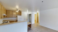 Unit #204 at 3630 6th Avenue in San Diego offers a practical, single-floor layout designed for comfortable everyday living. The open-concept design connects the kitchen directly to the main living area, creating a cohesive and functional flow throughout the space.

The kitchen features light wood cabinetry, a white refrigerator, and a peninsula counter with a laminate surface that extends into a breakfast bar area, providing additional seating and workspace. A ceiling fan with integrated lighting illuminates the kitchen, while flush-mount ceiling lights carry brightness into the adjoining living space.

The living area is finished with neutral-toned carpet, contributing to a warm and understated atmosphere. White walls throughout keep the interior feeling bright and clean. A built-in closet with a hanging rod is conveniently positioned off the main living area, offering accessible storage without occupying additional floor space.

Toward the rear of the unit, a full bathroom is visible, finished with light-toned flooring and wood-accented cabinetry, maintaining the warm, neutral palette consistent across the space. The textured ceiling in the kitchen zone adds a subtle architectural detail to the otherwise streamlined interior.