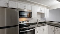 This single-floor unit offers a thoughtfully updated interior with a clean, neutral aesthetic throughout. The kitchen is a standout feature, showcasing crisp white shaker-style cabinetry, dark granite countertops, and a full suite of stainless steel appliances including a refrigerator, electric range, over-the-range microwave, and a stainless undermount sink. A tiled ceiling adds a classic touch, while light wood-look flooring creates a seamless transition into the adjacent living areas. The open layout connects the kitchen to a spacious living area, which is bathed in natural light from a window and features the same warm wood-look flooring, neutral gray walls, and a baseboard heating system. The bedroom is generously sized, offering ample room and a similar palette of soft gray tones, wood flooring, and a tiled ceiling. The bathroom is clean and functional, featuring a full tub-shower combination with white tile surround, wood flooring, and simple, modern fixtures. Throughout the unit, consistent finishes — from the white trim and neutral walls to the cohesive flooring — create a unified and move-in-ready environment that is both practical and visually appealing.