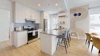 Located at 975 Avenue St-Charles in Saint-Lambert, Quebec, this well-designed 2-bedroom, 2-bathroom residence offers a thoughtfully laid-out single-floor plan with modern finishes throughout. Light hardwood flooring runs continuously across the space, complementing the bright, neutral palette that defines the overall aesthetic.

The open-concept living, dining, and kitchen area forms the heart of the home. The kitchen is equipped with white cabinetry, stainless steel appliances, a grey tile backsplash, and a generous island with bar seating — ideal for both meal preparation and casual dining. The dining area benefits from large windows that invite natural light, while the living room features a striking decorative accent wall with bold artistic wallpaper and warm sconce lighting.

The primary bedroom is a comfortable, well-proportioned retreat with its own closet access. The second bedroom also offers good natural light, a closet, and a wall-mounted air conditioning unit. Both bathrooms are finished with clean, contemporary materials — one featuring a glass-enclosed walk-in shower, wood-toned cabinetry, and tile flooring.

Additional highlights include a dedicated laundry room with side-by-side washer and dryer, three closets for ample storage, a welcoming entrance, and access to a private balcony. The space combines functional design with modern style in a well-connected Saint-Lambert location.