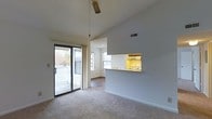 This single-floor residence offers an open, airy layout defined by soaring vaulted ceilings that lend a sense of spaciousness to the main living area. Soft, neutral-toned carpeting runs throughout, complementing the clean white walls and creating a warm, welcoming ambiance. A ceiling fan is centrally positioned beneath the high roofline, providing both function and a classic touch.

The living area flows naturally toward a set of sliding glass doors that open directly onto an outdoor deck, allowing for abundant natural light and easy access to the exterior. A secondary window nearby further brightens the space, offering views of the surrounding greenery.

The kitchen features a pass-through counter that connects it to the living area, promoting an open and communicative layout. Under-cabinet lighting illuminates the workspace, while the kitchen's positioning allows the cook to remain engaged with the rest of the room. A recessed ceiling light keeps the kitchen well-lit throughout the day.

Overall, the space presents a practical yet comfortable single-floor living environment, characterized by its vaulted ceilings, natural light, indoor-outdoor connectivity, and cohesive neutral palette — making it a functional and inviting place to call home.
