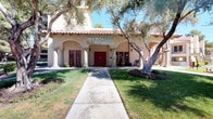 This single-story residential care facility is designed in a warm Spanish Colonial Revival style, characterized by stucco exterior walls, a red clay tile roof, arched colonnades, and rich red-painted doors and window trim. The inviting front entrance is framed by mature olive trees and lush green lawns, with a straight walkway leading to the main entry. A welcoming banner proudly displays the facility's commitment to its residents, setting a caring and community-oriented tone throughout.

The covered outdoor corridor features exposed wood beam ceilings, terracotta tile flooring, and a series of graceful arched openings supported by cylindrical columns. This shaded arcade serves as a transitional gathering space, furnished with ornate white wrought iron patio tables and chairs, offering residents a comfortable place to relax and socialize. Wall-mounted lantern-style light fixtures add a classic decorative touch. A vending machine is conveniently accessible along the corridor, adjacent to red-framed glass doors that lead to interior spaces.

The courtyard visible through the arches provides an open-air retreat with additional seating and landscaping, reinforcing the facility's emphasis on accessible outdoor living. The overall architectural design balances aesthetic charm with functional, resident-friendly amenities.