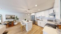 Located at 10 Arbutus Path in Mount Sinai, NY, this single-floor, 2-bedroom, 2-bathroom home offers a clean, modern aesthetic throughout. The open-concept kitchen is a standout, featuring crisp white shaker cabinetry, a marble-look tile backsplash, quartz countertops, stainless steel appliances, and a central island with seating — all illuminated by stylish pendant lighting and recessed fixtures. Light oak hardwood flooring flows through the kitchen and living areas, lending warmth to the bright, airy layout. The living room is comfortable and well-lit, with multiple windows allowing ample natural light. The generously sized primary bedroom includes plush carpeting and access to a private en-suite bathroom finished with large-format marble-look tile, a walk-in shower, and a sleek vanity with an LED-lit mirror. The second bedroom, also carpeted, offers a window with outdoor views and flexible use as a guest room or home office. The second bathroom features a full tub-shower combo clad in elegant marble-look tile, a white vanity, and coordinating tile flooring. Additional highlights include multiple closets, a dedicated laundry area, a utility room, and a hallway connecting all spaces efficiently. The home presents a cohesive, contemporary design throughout.