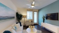 This single-floor residence offers a well-appointed living space that combines modern design with a warm, inviting atmosphere. The living area is anchored by rich, dark hardwood flooring that contrasts beautifully with soft, light-colored walls and a bold teal accent wall. A ceiling fan with integrated lighting is centrally positioned overhead, providing both comfort and ambient illumination. Large windows fitted with white horizontal blinds allow generous natural light to fill the room, creating an airy and open feel. A glass-paneled door leads directly to an outdoor area, further enhancing the sense of connectivity between interior and exterior spaces. The room is well-suited for comfortable living and entertaining, with a layout that accommodates seating and accent furniture with ease. Neutral tones throughout the main walls complement the vibrant accent wall, offering a balanced and contemporary aesthetic. The overall design strikes a blend of practicality and style, making it a functional yet visually engaging living environment.