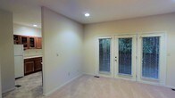 This single-floor space presents a clean, understated interior defined by neutral beige walls and plush carpeted flooring. The main living area is open and airy, benefiting from a recessed ceiling light that provides consistent overhead illumination. A set of white-framed French doors serves as a prominent architectural feature, fitted with horizontal blinds and offering a direct view of lush outdoor greenery, allowing natural light to filter into the room. The smooth white baseboards and trim throughout contribute to a polished, cohesive finish. Adjacent to the main living area, a partial view of a kitchen is visible, featuring warm wood-toned cabinetry that adds a subtle contrast to the lighter palette of the surrounding space. A floor vent is also present, indicating a central HVAC system. The overall ambiance is calm and versatile, with a simple, functional design that provides a neutral canvas well-suited for a variety of interior styles and furniture arrangements.