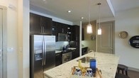 This modern single-floor residence showcases a thoughtfully designed interior that balances style and functionality. The kitchen is a standout centerpiece, featuring rich dark espresso cabinetry that extends from floor to ceiling, providing generous storage throughout. Stainless steel appliances, including a French door refrigerator with an in-door ice and water dispenser, a built-in microwave, and a range with oven, lend a polished, professional quality to the space. A light granite countertop island anchors the kitchen, offering ample prep space and a natural gathering point. The island is illuminated by a pair of elegant pendant lights with brushed nickel hardware, adding warmth and visual interest overhead. Recessed lighting throughout ensures the space is well-lit. A glass tile backsplash in cool tones adds texture and contrast against the dark cabinetry, while the under-counter wine cooler adds a refined touch. The open layout connects the kitchen to adjacent living areas, with warm neutral wall tones creating a cohesive and inviting ambiance throughout. The overall design presents a contemporary aesthetic with high-end finishes that elevate everyday living.