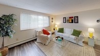 This single-floor unit offers a practical and cohesive layout designed for comfortable everyday living. The kitchen is a standout feature, outfitted with rich dark cabinetry, granite-look countertops, and a full suite of stainless steel appliances including a refrigerator, electric range, and built-in microwave. A double-basin sink and ample upper and lower cabinet storage make the kitchen both functional and visually striking. A breakfast bar with seating connects the kitchen to the open-concept living and dining area, promoting a sense of flow and connectivity throughout the space. The living area is generously sized, featuring wood-look plank flooring that extends throughout the unit, neutral-toned walls, and a large window fitted with vertical blinds that allows natural light to fill the room. The bedroom is a quiet, well-proportioned room with its own window for natural light, baseboard heating, and the same consistent wood-look flooring. The full bathroom features a clean, white aesthetic with a combined tub and shower, a vanity with storage, a medicine cabinet mirror, and tile flooring. A drop ceiling with flush-mount lighting is present throughout, contributing to the unit's consistent, understated finish.