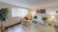 This single-floor unit offers a practical and cohesive layout designed for comfortable everyday living. The kitchen is a standout feature, outfitted with rich dark cabinetry, granite-look countertops, and a full suite of stainless steel appliances including a refrigerator, electric range, and built-in microwave. A double-basin sink and ample upper and lower cabinet storage make the kitchen both functional and visually striking. A breakfast bar with seating connects the kitchen to the open-concept living and dining area, promoting a sense of flow and connectivity throughout the space. The living area is generously sized, featuring wood-look plank flooring that extends throughout the unit, neutral-toned walls, and a large window fitted with vertical blinds that allows natural light to fill the room. The bedroom is a quiet, well-proportioned room with its own window for natural light, baseboard heating, and the same consistent wood-look flooring. The full bathroom features a clean, white aesthetic with a combined tub and shower, a vanity with storage, a medicine cabinet mirror, and tile flooring. A drop ceiling with flush-mount lighting is present throughout, contributing to the unit's consistent, understated finish.