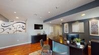 This single-floor professional office space combines contemporary design with warm, inviting elements to create a polished and functional environment. Rich hardwood flooring flows throughout the entire space, anchoring each area with a sense of warmth and cohesion.

The reception area serves as an impressive first impression, featuring a curved front desk, a wall-mounted flat-screen TV, and a bold decorative mural of sweeping floral and scroll motifs that extends across a prominent accent wall. Slate-blue architectural beams add a modern, structural contrast to the crisp white ceilings outfitted with recessed lighting.

Adjacent to the reception, a spacious lounge area offers comfortable seating, including a tufted gray sofa and vibrant red accent chairs, arranged around a glass-topped coffee table. A large window dressed with draping curtains allows natural light to fill the room, creating an open and welcoming atmosphere.

The breakroom is compact yet well-equipped, featuring white cabinetry, open shelving, a stainless steel refrigerator, and countertop appliances — providing staff with all essential amenities. A workstation area with built-in counters and frosted glass cabinetry adds additional functionality to the space.

Overall, the office balances professional utility with thoughtful design details, making it well-suited for client-facing operations.