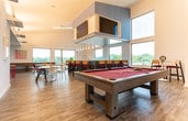 Located in Atlanta, GA (30316), this contemporary community amenity lounge is designed to offer a dynamic and social atmosphere across a single, open-plan floor. The space is characterized by soaring double-height ceilings, light wood-style flooring, and an abundance of natural light streaming through large floor-to-ceiling windows that frame sweeping views of the surrounding cityscape and greenery.

The main lounge area is thoughtfully divided into distinct activity zones. A full-size pool table anchors the central game area, complemented by a regulation shuffleboard table positioned near expansive windows. An arcade gaming corner adds a playful touch, equipped with a mounted television and a classic arcade cabinet. Colorful artwork punctuates the white walls throughout, adding personality and vibrancy to the space.

A sleek bar and kitchenette serves as the social hub, featuring herringbone-patterned tile detailing, modern gray cabinetry, marble-look countertops, a wine refrigerator, and elegant pendant lighting. High-top bar seating with red cushions and woven-back chairs lines the counter, creating an inviting spot for casual gatherings. Additional lounge seating, including a plush sofa and eclectic round café tables with mixed chairs, provides comfortable spaces for relaxation and conversation throughout the room.