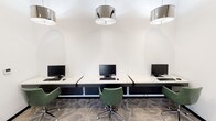 Located at 3503 W Walnut Hill Lane in Irving, Texas, this single-floor business center offers a polished and thoughtfully designed environment suited for productivity and comfort. The space is anchored by a well-appointed computer lounge featuring multiple workstations set along marble-topped desks with dark bases, complemented by ergonomic green upholstered chairs. Polished chrome drum pendant lights hang overhead, casting a clean, bright light across the patterned carpet flooring that adds texture and visual depth throughout. The workstation area is divided into two sections, accommodating both individual and side-by-side work arrangements, making the space versatile for various needs. A welcoming lounge area greets visitors near the entrance, furnished with a tufted linen settee accented by emerald green throw pillows. The walls serve as a curated gallery, showcasing an eclectic collection of modern and abstract artwork in varied sizes and frames, lending the space a creative, sophisticated ambiance. A glass-paneled entry door with black framing allows natural light to filter in and offers views of the landscaped exterior. Dark-framed double glass doors connect to an adjacent corridor, maintaining an open and airy flow. The overall aesthetic balances modern design with warm, inviting touches, creating a professional yet comfortable setting.