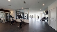 Located at 1449 S Church St in Charlotte's South End neighborhood, this well-appointed fitness center offers a spacious, single-floor layout designed to accommodate a full range of workout needs. The space features a clean, contemporary design with light-colored walls, recessed lighting, and dark rubber flooring that provides both durability and comfort underfoot. Large floor-to-ceiling windows line multiple walls, flooding the interior with abundant natural light and offering views of the surrounding outdoor area, including a glimpsed courtyard or pool deck. The cardio zone is well-equipped with treadmills, stationary spin bikes with integrated display screens, and elliptical machines. A dedicated strength training area includes a multi-station cable machine, a variety of selectorized machines for targeted muscle groups, a full dumbbell rack, and a functional training rig complete with a punching bag, battle ropes, medicine balls, and a trampoline. Floor-to-ceiling mirrors in the strength zone enhance the sense of space and allow for proper form checks. A water fountain station is conveniently positioned near a glass door leading to the outdoor area. The overall ambiance is bright, professional, and energizing, making this facility a comprehensive resource for fitness enthusiasts.