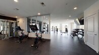 Located at 1449 S Church St in Charlotte's South End neighborhood, this well-appointed fitness center offers a spacious, single-floor layout designed to accommodate a full range of workout needs. The space features a clean, contemporary design with light-colored walls, recessed lighting, and dark rubber flooring that provides both durability and comfort underfoot. Large floor-to-ceiling windows line multiple walls, flooding the interior with abundant natural light and offering views of the surrounding outdoor area, including a glimpsed courtyard or pool deck. The cardio zone is well-equipped with treadmills, stationary spin bikes with integrated display screens, and elliptical machines. A dedicated strength training area includes a multi-station cable machine, a variety of selectorized machines for targeted muscle groups, a full dumbbell rack, and a functional training rig complete with a punching bag, battle ropes, medicine balls, and a trampoline. Floor-to-ceiling mirrors in the strength zone enhance the sense of space and allow for proper form checks. A water fountain station is conveniently positioned near a glass door leading to the outdoor area. The overall ambiance is bright, professional, and energizing, making this facility a comprehensive resource for fitness enthusiasts.