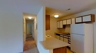 This single-floor residence offers a practical and comfortable layout designed for everyday living. The kitchen is a standout feature, equipped with warm wood cabinetry in rich brown tones paired with white upper cabinets, providing ample storage space. A white refrigerator and built-in stovetop are complemented by a double sink set against a dark countertop. A peninsula with a light-colored countertop extends into the adjacent space, creating a convenient breakfast bar that connects the kitchen to the main living area. Flush-mount ceiling lighting illuminates both the kitchen and hallway with a warm, inviting glow. A central hallway stretches from the kitchen area, lined with smooth white walls and carpeted flooring in a neutral tone that continues throughout the residence. Doors along the hallway lead to additional rooms, suggesting a multi-room floor plan suited for comfortable daily use. The overall ambiance is warm and understated, with a classic residential aesthetic defined by neutral tones, functional cabinetry, and a cohesive layout that maximizes the use of available space on a single level.