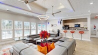Located at 1038 South Hairston Road in Stone Mountain, Georgia, this single-floor space offers a well-appointed, open-plan interior designed to serve both collaborative and functional purposes. The main living area is expansive and bright, with light-toned hardwood flooring running throughout and crisp white walls that enhance the sense of openness. A tiled fireplace with a wall-mounted flat-screen TV above it anchors one end of the space, while large windows fitted with white horizontal blinds flood the room with natural light and offer views of the surrounding greenery and an outdoor pool area. French doors provide direct access to the pool-facing exterior.

The open layout seamlessly integrates a lounge area, furnished with a generous sectional sofa and a glass-and-chrome coffee table atop a bold area rug, alongside a dedicated workspace equipped with multiple desks, ergonomic chairs, computers, and office equipment. Vibrant abstract artwork adorns the walls, adding color and creative energy to the environment. A kitchenette with bar-height seating and orange accent stools is tucked toward the rear, complemented by a ceiling fan and recessed lighting overhead. Crown molding and a textured ceiling add architectural refinement throughout. Ambient blue accent lighting contributes a contemporary, polished atmosphere.