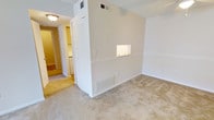 This single-floor residence offers a straightforward and functional layout designed for comfortable everyday living. The main living area is finished with neutral wall-to-wall carpeting that lends a warm, understated feel to the space. A defining architectural detail is the built-in pass-through window that connects the living area to an adjacent room, allowing light to flow between spaces and adding a practical touch to the open concept design.

The walls are painted in a clean, neutral tone throughout, contributing to a bright and cohesive atmosphere. A louvered closet door provides convenient built-in storage directly off the main living area, keeping essentials neatly out of sight. HVAC vents are integrated into both the walls and ceiling, ensuring efficient climate control throughout the unit.

A hallway connects the main living space to additional rooms, with a glimpse of what appears to be a laundry or utility area visible in the background, complete with white appliances. The ceiling-mounted light fixture provides consistent, even illumination. Overall, the space presents a clean, move-in ready environment with a practical design that prioritizes functionality and ease of living across its single level.