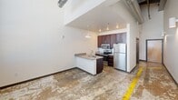 This efficiently designed unit in Burlington, NC showcases an industrial-chic aesthetic with soaring ceilings, exposed ductwork, and polished concrete floors accented by original brick walls. The open-concept living and kitchen area features stainless steel appliances, rich dark cabinetry, granite countertops, and a peninsula with a built-in sink — all illuminated by a pendant light and recessed lighting. The living area offers wall-mounted sconce lighting and generous vertical space. A full bathroom includes a tub/shower combo with a curved curtain rod, and a dedicated laundry nook houses a stacked washer and dryer alongside built-in wire shelving for added storage.