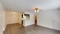 This single-floor residence offers a thoughtfully laid-out living space designed for both comfort and functionality. The entry is accessible via a covered patio, framed by a decorative wrought iron security door and wood-panel fencing, with pleasant views of mature trees and a well-maintained courtyard beyond.

Inside, the main living area is open and airy, featuring warm wood-look laminate flooring that extends throughout the space. A textured popcorn ceiling adds classic character, while a ceiling fan with integrated lighting provides both comfort and illumination. A pendant light near the dining area enhances the warm ambiance of the open-plan layout.

The kitchen is positioned at the heart of the home, separated from the living area by a raised pass-through counter that allows for easy interaction between spaces. It features dark cabinetry, white appliances, and ample counter space for everyday use. The kitchen opens conveniently toward an adjacent hallway that leads further into the home.

Natural light filters in through the front-facing windows and patio, creating a bright and inviting atmosphere throughout. The overall design reflects a practical, comfortable layout well-suited for everyday living, with a seamless flow between the outdoor entry, main living space, and kitchen.