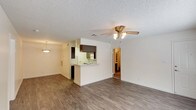 This single-floor residence offers a thoughtfully laid-out living space designed for both comfort and functionality. The entry is accessible via a covered patio, framed by a decorative wrought iron security door and wood-panel fencing, with pleasant views of mature trees and a well-maintained courtyard beyond.

Inside, the main living area is open and airy, featuring warm wood-look laminate flooring that extends throughout the space. A textured popcorn ceiling adds classic character, while a ceiling fan with integrated lighting provides both comfort and illumination. A pendant light near the dining area enhances the warm ambiance of the open-plan layout.

The kitchen is positioned at the heart of the home, separated from the living area by a raised pass-through counter that allows for easy interaction between spaces. It features dark cabinetry, white appliances, and ample counter space for everyday use. The kitchen opens conveniently toward an adjacent hallway that leads further into the home.

Natural light filters in through the front-facing windows and patio, creating a bright and inviting atmosphere throughout. The overall design reflects a practical, comfortable layout well-suited for everyday living, with a seamless flow between the outdoor entry, main living space, and kitchen.
