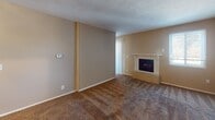 This single-floor unit offers a straightforward, functional layout connecting several well-defined spaces. The living room serves as the central gathering area, finished with wall-to-wall carpeting in a warm brown tone and neutral beige walls. A fireplace with a light-toned surround and built-in cabinetry below serves as the room's focal point, while vertical blinds on an adjacent window allow for natural light control. A textured ceiling adds a classic residential character throughout the space. The entry area opens directly into the living room, providing a seamless transition from the front door to the main living space, with the kitchen visible from this vantage point.

The galley-style kitchen is efficiently designed, featuring rich wood cabinetry lining both sides, dark granite-style countertops, a double sink, and black appliances including an electric range and refrigerator. Tile flooring and a flush-mounted ceiling light complete the functional aesthetic.

A carpeted hallway connects the living areas to additional rooms, lined with white sliding closet doors that offer generous built-in storage. The overall color palette remains consistent throughout—warm beige tones, white trim, and brown carpet—creating a cohesive and understated ambiance.