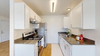 This single-story, 2-bedroom, 1-bathroom unit offers a clean, modern aesthetic with a practical and efficient layout. Warm hardwood-style flooring runs throughout the entire space, creating a cohesive and inviting atmosphere.

The living area is bright and open, featuring large windows that admit generous natural light and offer views of the surrounding neighborhood. The space is finished with crisp white walls and a textured ceiling, contributing to a fresh, airy feel.

The galley-style kitchen is well-appointed, featuring white shaker cabinetry, dark countertops, and stainless steel appliances including a range and refrigerator. Overhead lighting ensures the kitchen is bright and functional, while ample cabinet storage lines both sides of the corridor layout.

Both bedrooms are comfortably sized, each offering sliding-door closets with built-in rods for storage. The primary bedroom provides a generous footprint, while the second bedroom benefits from a window that brings in natural light. Both rooms are finished with the same neutral palette found throughout the unit.

The bathroom is neatly designed, featuring a white vanity with a dark countertop, an undermount sink with modern fixtures, a large mirror, and a shower with a curtain rod. Baseboard heating is present in the bedrooms and bathroom, ensuring year-round comfort. A hallway connects the rooms, completing this efficient and well-maintained single-floor layout.