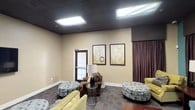 This single-floor lounge space is designed to accommodate comfortable gatherings with a carefully curated selection of furnishings and a consistent interior palette. The room features a dark drop-tile ceiling with recessed fluorescent panel lighting that evenly illuminates the space, contrasting with the warm beige and teal-accented walls. Rich, floor-length chocolate brown drapery lines multiple walls, adding depth and a formal touch to the overall aesthetic.

The seating arrangement is varied and plentiful, incorporating a light blue upholstered sofa adorned with decorative throw pillows, chartreuse accent chairs, a brown leather armchair, and several round tufted ottomans in a complementary spotted fabric. A glass-top side table with a sculptural metal base and table lamps with black ceramic bases are distributed throughout, enhancing both functionality and ambiance. A wooden coffee table anchors the central seating area.

Patterned area carpeting in muted tones of blue, gray, and beige covers most of the floor, grounding the eclectic furniture groupings. Framed abstract wall art and a wall-mounted flat-screen television contribute to the finished, intentional decor. A windowed door on one wall provides a modest connection to natural light, while an adjacent room visible through a doorway suggests additional space beyond.