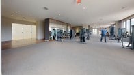 This well-appointed fitness and recreation facility spans a single floor and is thoughtfully organized into distinct zones for exercise and leisure. The main gym area is expansive and flooded with natural light from large floor-to-ceiling windows that frame outdoor views, creating an energizing atmosphere. A row of treadmills and cardio machines lines the windowed perimeter, while a comprehensive selection of strength training equipment — including cable machines, weight stations, and resistance machines — is arranged throughout the space. Mirrored wall panels framed in warm wood accents enhance the sense of openness and allow users to monitor their form. A dedicated flexibility and wellness zone houses stability balls, yoga mats, and resistance accessories, providing a well-rounded fitness experience. Adjacent to the main gym is a spacious open-floor studio area with light hardwood flooring, accent artwork, and a built-in bench, ideal for yoga, stretching, or group classes. A separate children's activity room rounds out the facility, featuring light wood floors, a bold striped accent wall, a large flat-screen television, circular bookshelves stocked with books, activity tables, and a plush area rug — offering a bright, playful environment for younger guests.